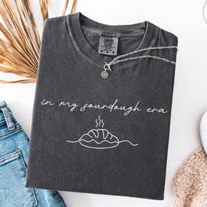 Pode incluir: T-shirt cinza escuro com o texto "in my sourdough era" e um gr&aacute;fico de p&atilde;o. Um colar de prata com um pingente de sol est&aacute; na camisa. A camisa est&aacute; dobrada em uma superf&iacute;cie branca ao lado de jeans.
