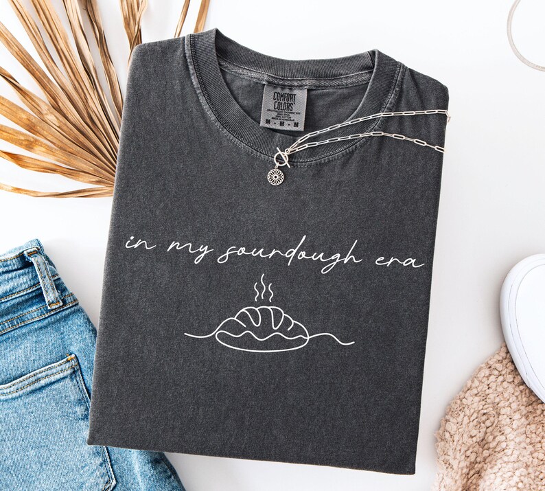 Pode incluir: T-shirt cinza escuro com o texto "in my sourdough era" e um gr&aacute;fico de p&atilde;o. Um colar de prata com um pingente de sol est&aacute; na camisa. A camisa est&aacute; dobrada em uma superf&iacute;cie branca ao lado de jeans.