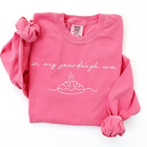 Pode incluir: Uma camisola rosa de manga comprida com a frase "in my sourdough era" em it&aacute;lico. Abaixo do texto, um desenho a tra&ccedil;o branco de um p&atilde;o com vapor. A camisola tem gola redonda e punhos enrolados.