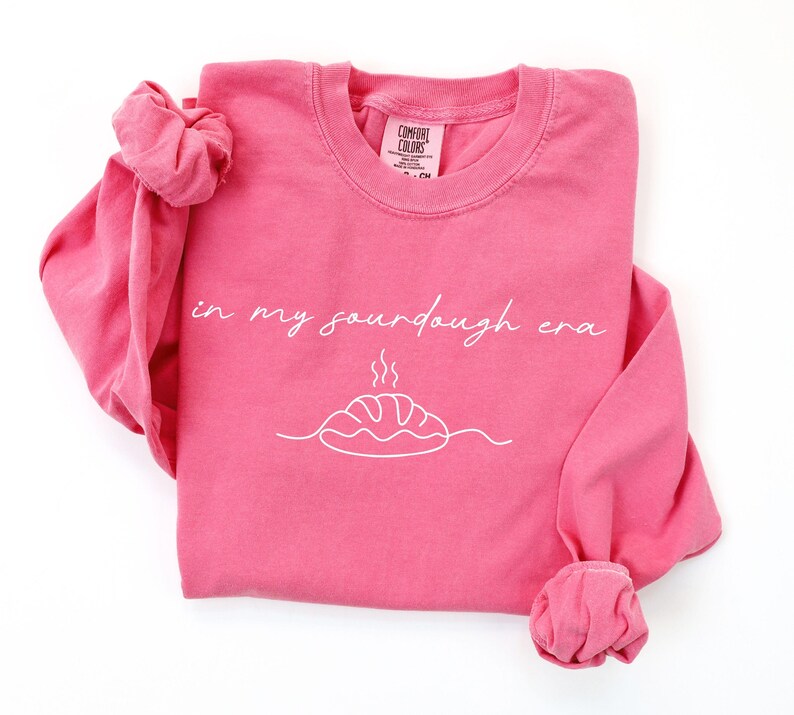 Pode incluir: Uma camisola rosa de manga comprida com a frase "in my sourdough era" em it&aacute;lico. Abaixo do texto, um desenho a tra&ccedil;o branco de um p&atilde;o com vapor. A camisola tem gola redonda e punhos enrolados.