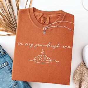 Pode incluir: Uma camiseta cor de ferrugem com a frase "in my sourdough era" em escrita branca, juntamente com um gr&aacute;fico de p&atilde;o. Um colar de prata com um pingente de sol repousa sobre a camisa. A camisa est&aacute; dobrada sobre uma superf&iacute;cie branca ao lado de jeans azuis.