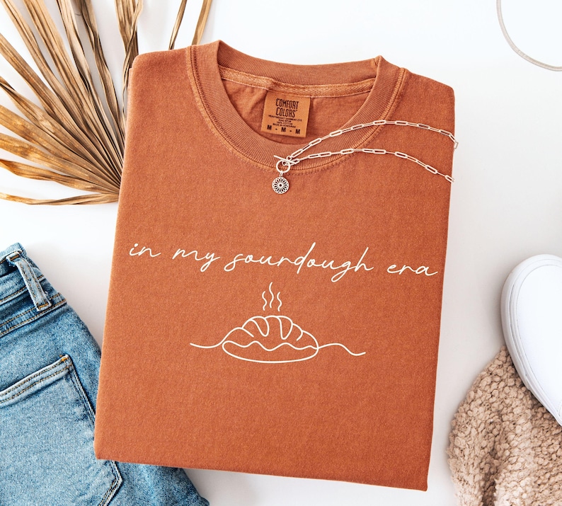 Pode incluir: Uma camiseta cor de ferrugem com a frase "in my sourdough era" em escrita branca, juntamente com um gr&aacute;fico de p&atilde;o. Um colar de prata com um pingente de sol repousa sobre a camisa. A camisa est&aacute; dobrada sobre uma superf&iacute;cie branca ao lado de jeans azuis.