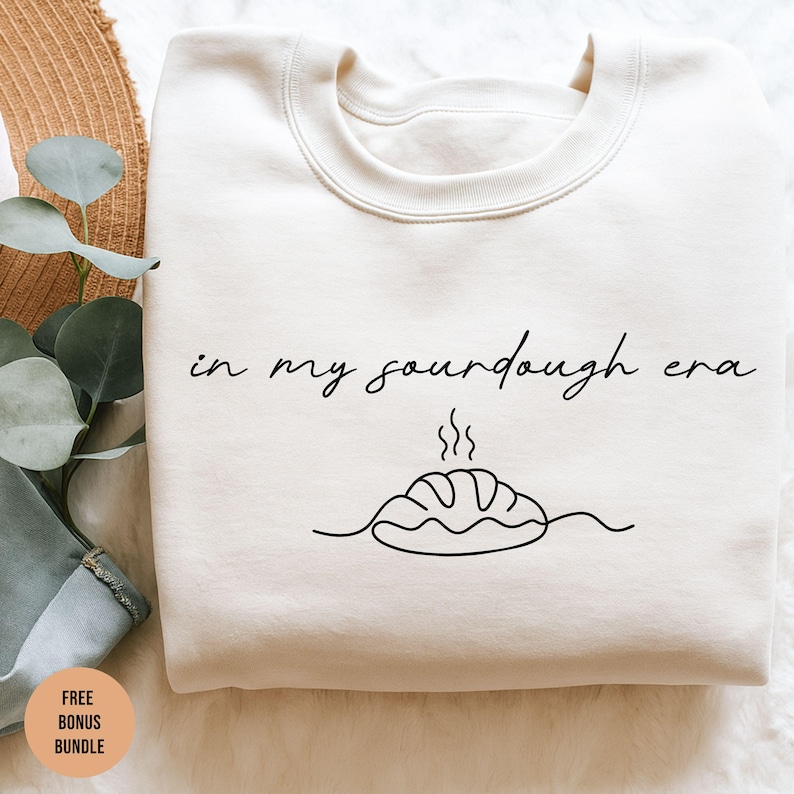Pode incluir: Camisola creme com a frase "in my sourdough era" em escrita preta. Abaixo do texto, um desenho de um p&atilde;o com vapor saindo. A camisola est&aacute; dobrada.