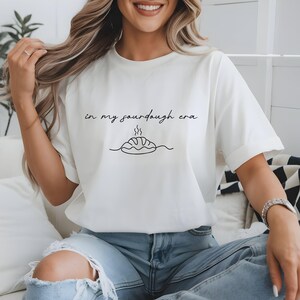 Pode incluir: Camiseta branca com a frase "in my sourdough era" em fonte manuscrita. Abaixo, um desenho de linha de um p&atilde;o com vapor. A camiseta &eacute; usada com jeans azuis.