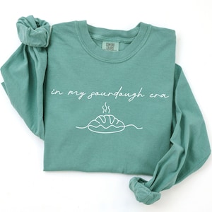 Pode incluir: Uma camisa de manga comprida verde-azulado com as palavras "in my sourdough era" em branco. Abaixo do texto, h&aacute; um desenho de linha branca de um p&atilde;o. A camisa &eacute; feita de um material macio.