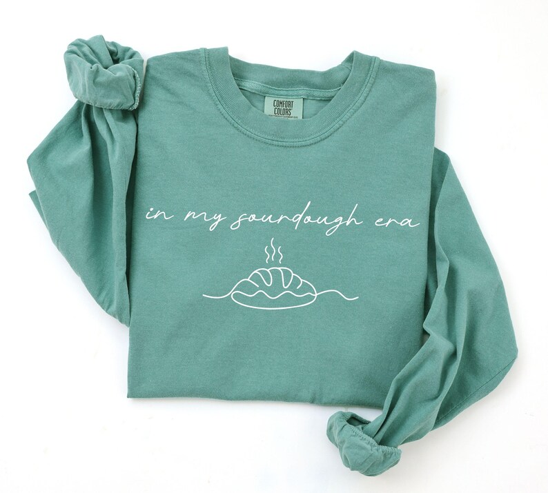 Pode incluir: Uma camisa de manga comprida verde-azulado com as palavras "in my sourdough era" em branco. Abaixo do texto, h&aacute; um desenho de linha branca de um p&atilde;o. A camisa &eacute; feita de um material macio.