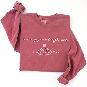 Pode incluir: Uma camisa de manga comprida bord&ocirc; com a frase "in my sourdough era" em escrita branca. Abaixo do texto, h&aacute; um desenho de linha branca de um p&atilde;o. A camisa tem gola redonda e mangas com punhos.