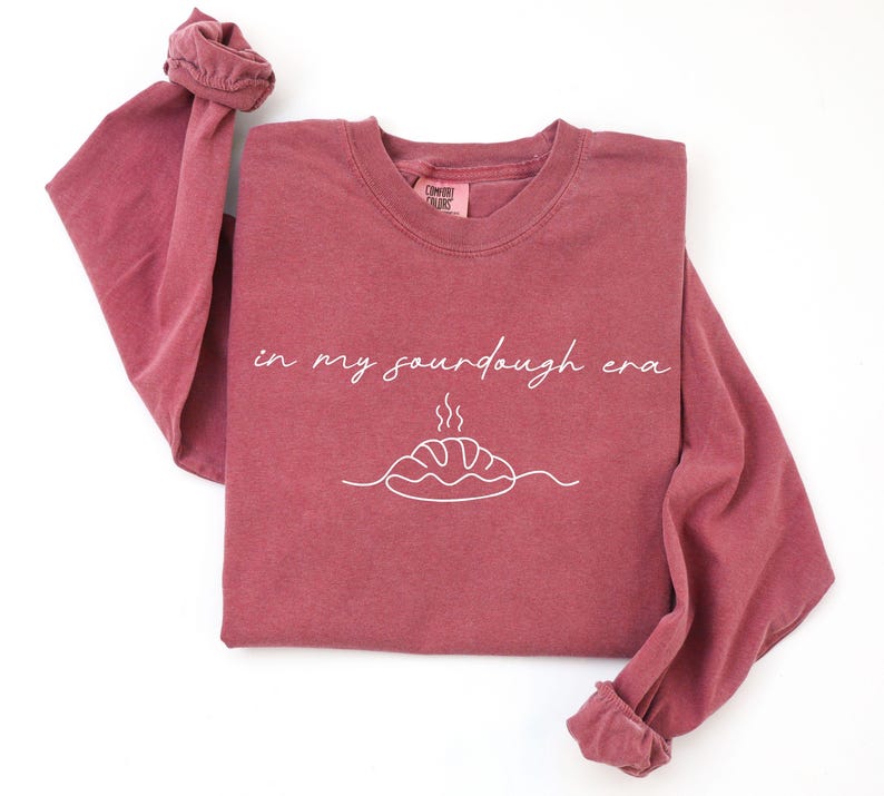 Pode incluir: Uma camisa de manga comprida bord&ocirc; com a frase "in my sourdough era" em escrita branca. Abaixo do texto, h&aacute; um desenho de linha branca de um p&atilde;o. A camisa tem gola redonda e mangas com punhos.