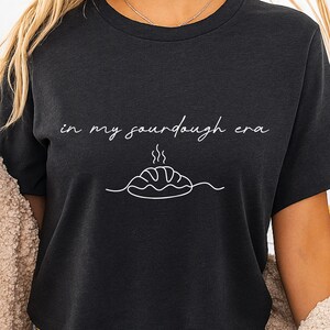 Pode incluir: Camiseta cinza escuro com a frase "in my sourdough era" em escrita branca sobre um desenho de linha de um p&atilde;o com vapor. A camiseta tem gola redonda e mangas curtas.