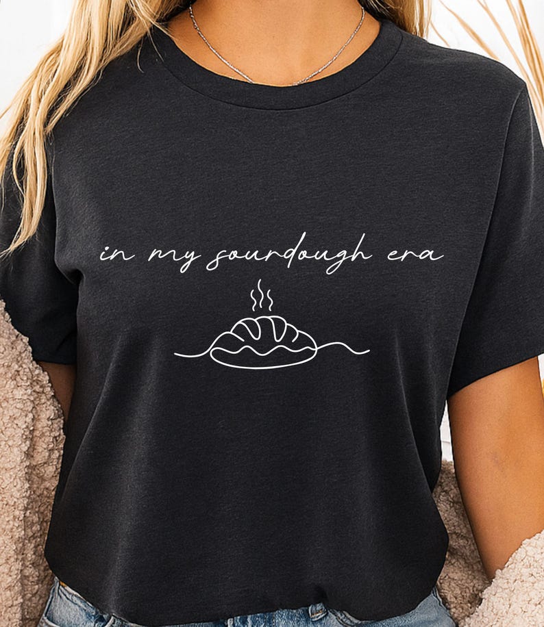 Pode incluir: Camiseta cinza escuro com a frase "in my sourdough era" em escrita branca sobre um desenho de linha de um p&atilde;o com vapor. A camiseta tem gola redonda e mangas curtas.