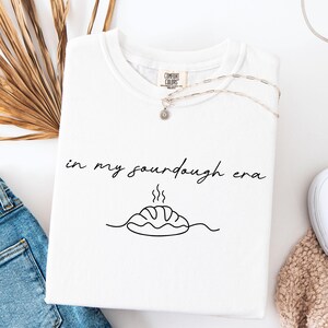 Pode incluir: Camiseta branca com a frase "in my sourdough era" em escrita preta. Abaixo do texto, h&aacute; um desenho de linha de um p&atilde;o com vapor. Um colar de prata com um pequeno pingente est&aacute; na camiseta.