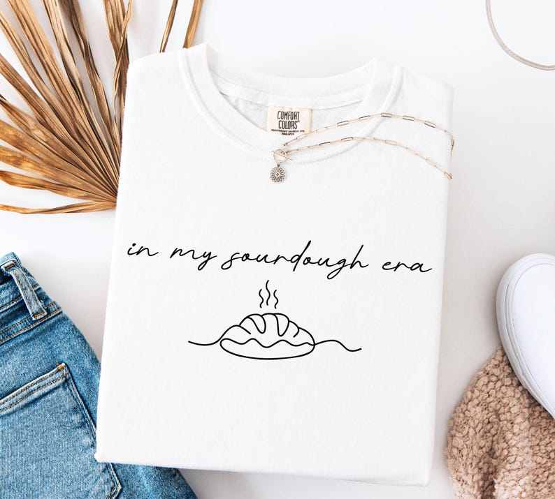 Pode incluir: Camiseta branca com a frase "in my sourdough era" em escrita preta. Abaixo do texto, h&aacute; um desenho de linha de um p&atilde;o com vapor. Um colar de prata com um pequeno pingente est&aacute; na camiseta.