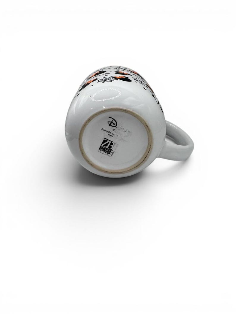 May include: A white ceramic mug featuring a Halloween-themed pattern with orange and black Minnie Mouse silhouettes, spiderwebs, and bats. The base displays the Disney logo, dishwasher safe text, and the Zrike Brands logo in black ink.