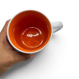 May include: A ceramic mug featuring a bright orange interior and a white exterior. The mug has a smooth, glossy finish on the inside and a sturdy handle attached to the side. The view is from directly above, showing the circular opening of the cup.