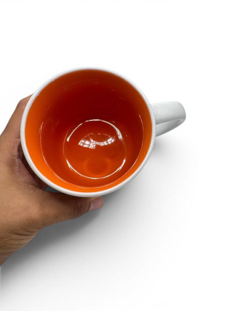 May include: A ceramic mug featuring a bright orange interior and a white exterior. The mug has a smooth, glossy finish on the inside and a sturdy handle attached to the side. The view is from directly above, showing the circular opening of the cup.