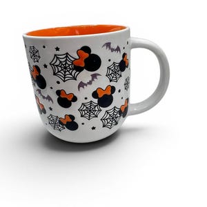 May include: A white ceramic mug with an orange interior featuring a repeating pattern of black mouse-ear silhouettes with orange bows, black spiderwebs, small stars, and grey bats scattered across the exterior surface and a curved handle.
