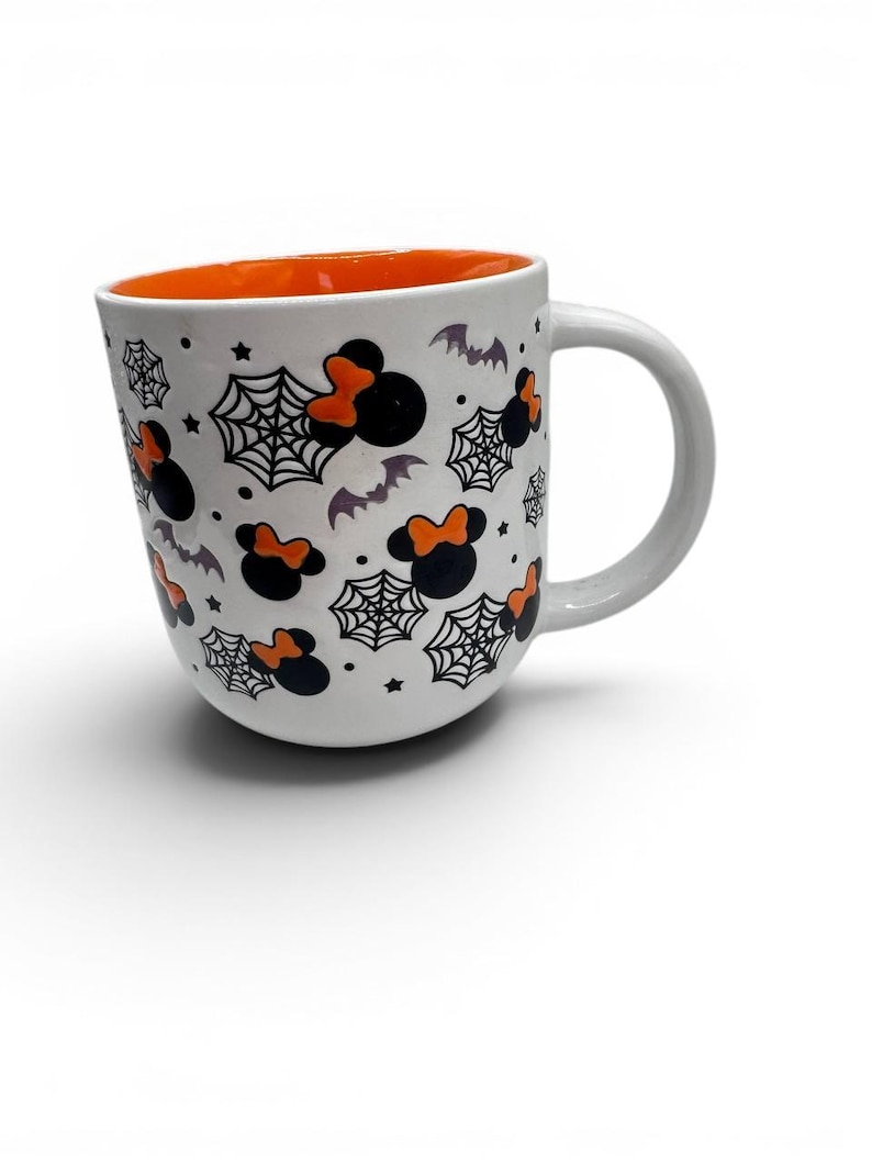 May include: A white ceramic mug with an orange interior featuring a repeating pattern of black mouse-ear silhouettes with orange bows, black spiderwebs, small stars, and grey bats scattered across the exterior surface and a curved handle.