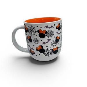 May include: A white ceramic mug with a rounded shape and a bright orange interior. The exterior features a repeating pattern of black mouse-ear silhouettes with orange bows, black spider webs, small purple bats, and tiny black stars.
