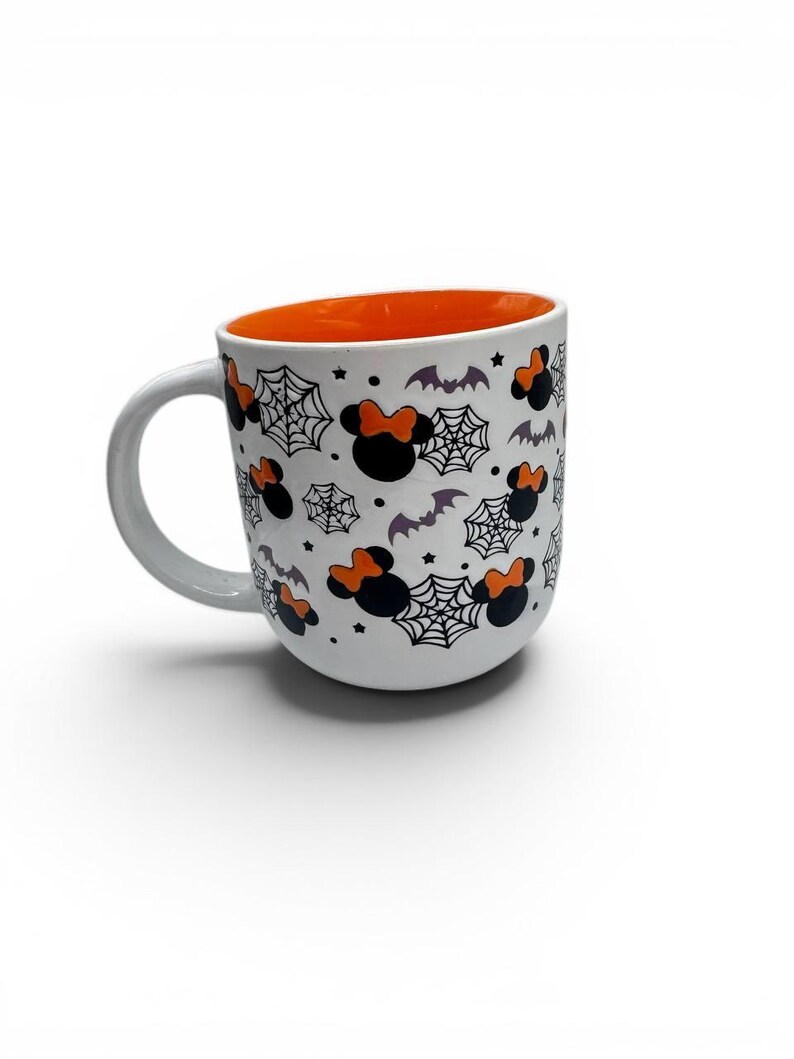 May include: A white ceramic mug with a rounded shape and a bright orange interior. The exterior features a repeating pattern of black mouse-ear silhouettes with orange bows, black spider webs, small purple bats, and tiny black stars.
