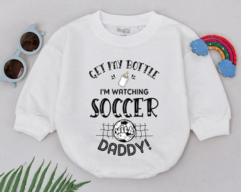 May include: White baby bodysuit with the text "Get My Bottle I'm Watching Soccer Daddy!" in black. The bodysuit features a soccer ball graphic and a bottle illustration. A rainbow and sunglasses are on the side.