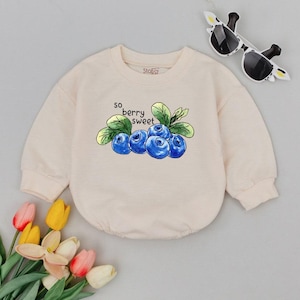 May include: A light beige baby sweatshirt with a crew neck and long sleeves. The front features a watercolor illustration of blueberries and the text "so berry sweet". Sunglasses are in the upper right corner.
