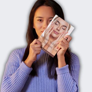 Puede incluir: Una mujer sostiene un libro titulado "Radiant Skin, Radiant You". La portada del libro presenta un primer plano de un rostro con manos tocando las mejillas. La mujer lleva un su&eacute;ter morado claro.