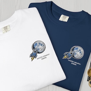 Puede incluir: Dos camisetas, una blanca y otra azul marino, cada una con un diseño bordado de un cohete y una luna. El texto "MOON Mission 2026" está debajo del diseño. También se ve una brújula y un mapa estelar.