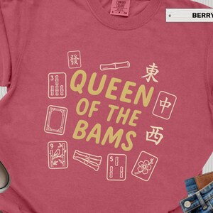 May include: Berry-colored t-shirt with the text "QUEEN OF THE BAMS" in gold, surrounded by white Mahjong tiles. The shirt is a Comfort Colors brand, with a tag indicating sizes S-2XL.