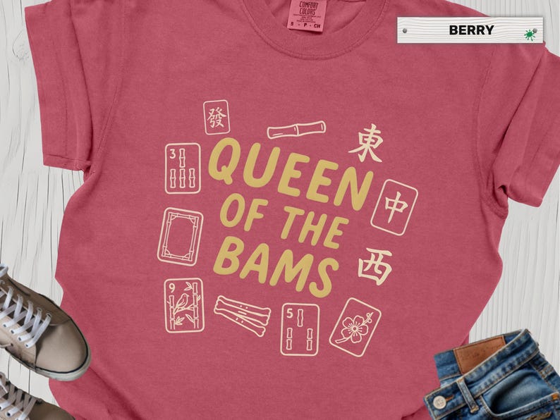 May include: Berry-colored t-shirt with the text "QUEEN OF THE BAMS" in gold, surrounded by white Mahjong tiles. The shirt is a Comfort Colors brand, with a tag indicating sizes S-2XL.