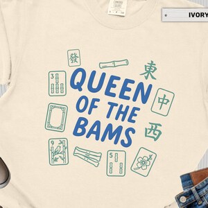 May include: Ivory-colored t-shirt with the text "QUEEN OF THE BAMS" in blue, surrounded by green Mahjong tiles. The shirt is made of a soft material and is suitable for casual wear.