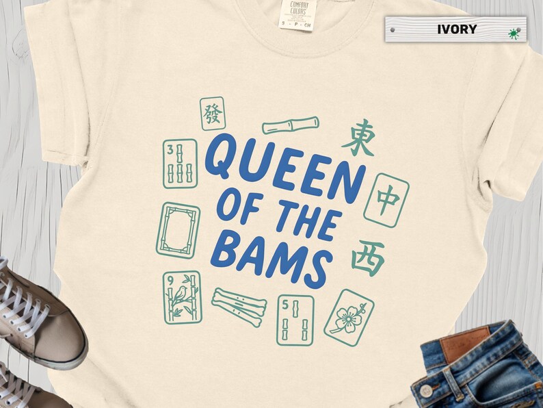 May include: Ivory-colored t-shirt with the text "QUEEN OF THE BAMS" in blue, surrounded by green Mahjong tiles. The shirt is made of a soft material and is suitable for casual wear.