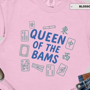 May include: A light pink t-shirt with the text "QUEEN OF THE BAMS" in blue, surrounded by green and blue Mahjong tiles. The word "BLOSSOM" is printed on a white label.