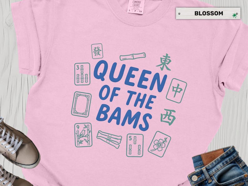 May include: A light pink t-shirt with the text "QUEEN OF THE BAMS" in blue, surrounded by green and blue Mahjong tiles. The word "BLOSSOM" is printed on a white label.