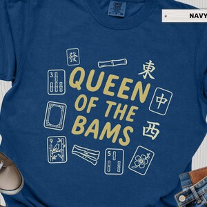 May include: Navy blue t-shirt with the text "QUEEN OF THE BAMS" in gold, surrounded by white and gold Mahjong tiles. The shirt is made of a soft material.