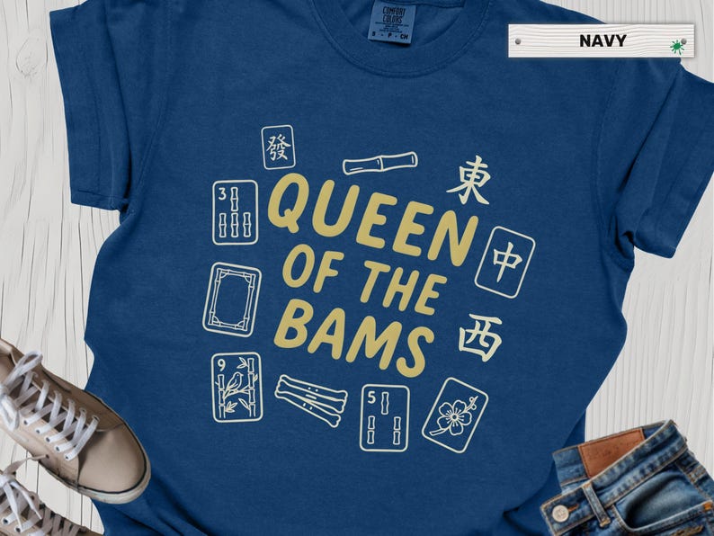 May include: Navy blue t-shirt with the text "QUEEN OF THE BAMS" in gold, surrounded by white and gold Mahjong tiles. The shirt is made of a soft material.