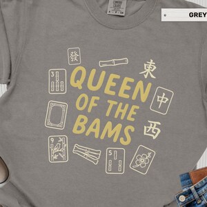 May include: A grey short-sleeved t-shirt featuring the text QUEEN OF THE BAMS in yellow lettering, surrounded by various Mahjong tile illustrations and Chinese characters printed in a light cream color on the front of the garment.