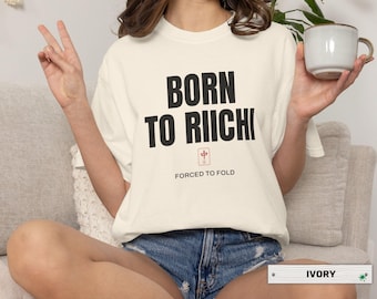 May include: Ivory-colored t-shirt with the text "BORN TO RIICHI" in bold black letters. Below the text is a red and white symbol, and the words "FORCED TO FOLD". The model is wearing denim shorts.
