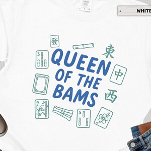 May include: A white t-shirt featuring the blue text QUEEN OF THE BAMS in the center, surrounded by green illustrations of mahjong tiles, bamboo stalks, and Chinese characters. The shirt has a crew neck and short sleeves.