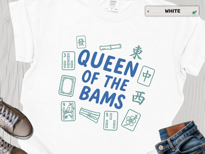 May include: A white t-shirt featuring the blue text QUEEN OF THE BAMS in the center, surrounded by green illustrations of mahjong tiles, bamboo stalks, and Chinese characters. The shirt has a crew neck and short sleeves.