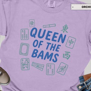 May include: Orchid-colored t-shirt with the text "QUEEN OF THE BAMS" in blue, surrounded by green Mahjong tiles and bamboo. The shirt is a crew neck style.
