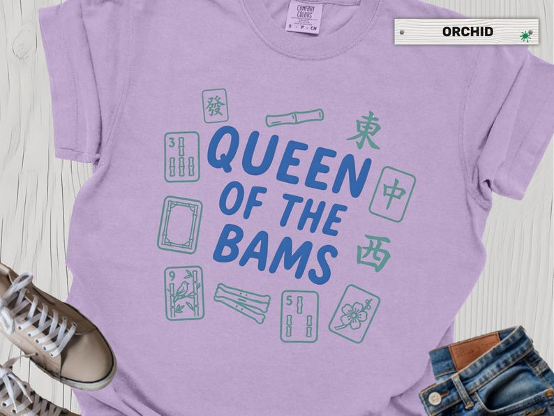 May include: Orchid-colored t-shirt with the text "QUEEN OF THE BAMS" in blue, surrounded by green Mahjong tiles and bamboo. The shirt is a crew neck style.