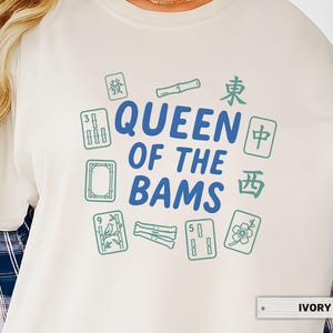 May include: Ivory-colored sweatshirt with the text "QUEEN OF THE BAMS" in blue, surrounded by green Mahjong tiles and Chinese characters. The shirt is a crew neck style.