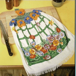 May include: A vintage book cover featuring a crocheted kitchen towel with a floral design. The towel has a green grid pattern and is adorned with orange, blue, and red flowers. The text "Kitchen Towels" is displayed at the top.