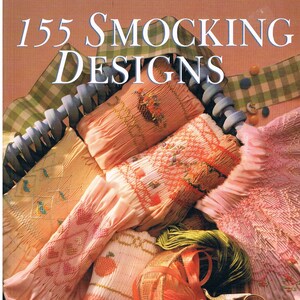 May include: The book cover reads "155 Smocking Designs" with examples of smocked fabric in peach, pink, and yellow. A basket contains thread, scissors, and pins. Green and blue ribbons are also visible.