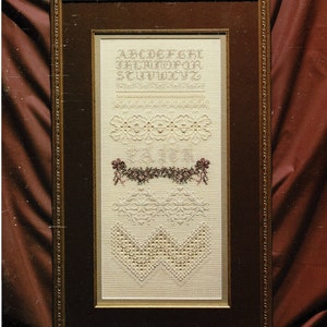 May include: A framed cross-stitch sampler with a brown wooden frame. The sampler features embroidered alphabets, decorative borders, and floral elements in cream and burgundy hues. The text "CROSS 'N PATCH" and "The Faith Sampler" are visible.