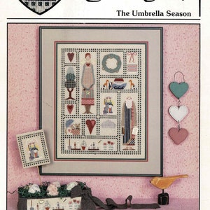 May include: A cross-stitch pattern featuring a charming house with a red roof and a blue door, surrounded by a heart-shaped frame. The pattern is titled "The Umbrella Season" and is part of the "HeartStrings" collection by Pat Thode.