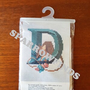 May include: A cross-stitch kit featuring the letter "D" with a floral design. The kit includes 100% cotton fabric, threads, and a needle. The kit is 21 cm x 20 cm and is made in Australia.