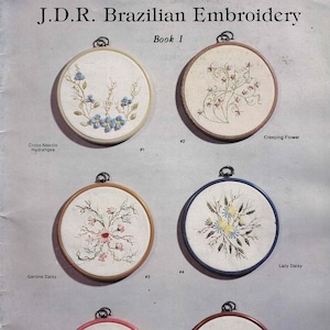 May include: A page from "J.D.R. Brazilian Embroidery Book I" featuring six embroidered floral designs in round hoops. Each hoop has a different floral pattern, including hydrangeas, daisies, and peach blossoms. The text includes the names of the designs and the author's name.