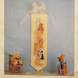 May include: A cross-stitch pattern featuring a stack of teddy bears with a bell pull. The pattern is on a cream-coloured fabric with a light blue background. The pattern is titled "Teddy Bears Bell Pull" and is designed by Donna Vermillion. The pattern is from the "Designer Series" by Sudbury House.