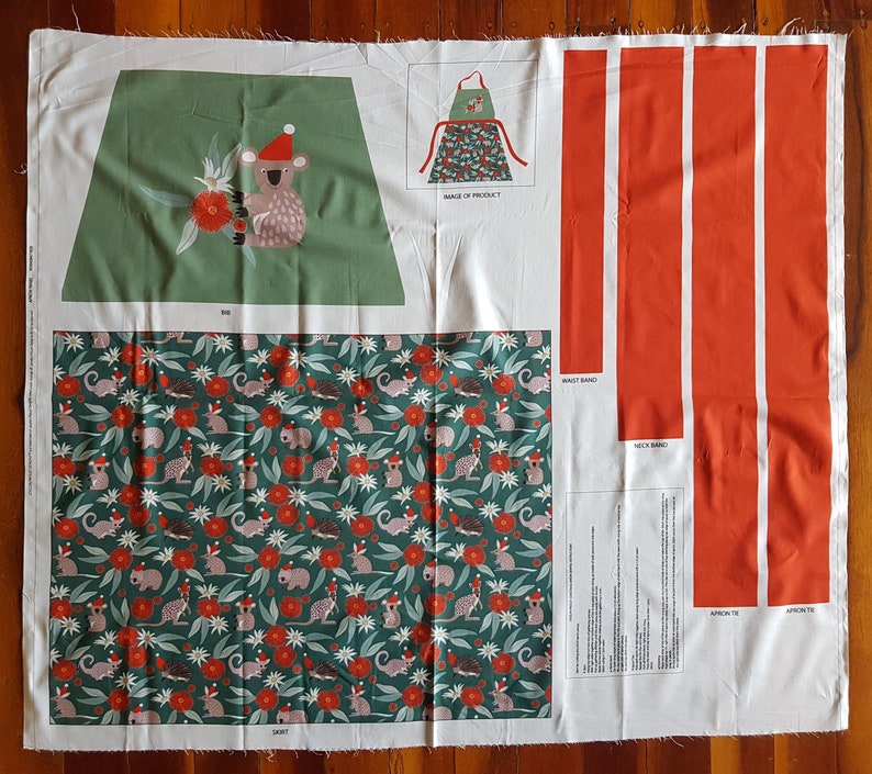 May include: A sewing pattern for a Christmas-themed apron featuring a koala wearing a Santa hat. The pattern includes instructions for the apron, neck band, waist band, and apron tie. The fabric is a green and red print with koalas, flowers, and leaves.